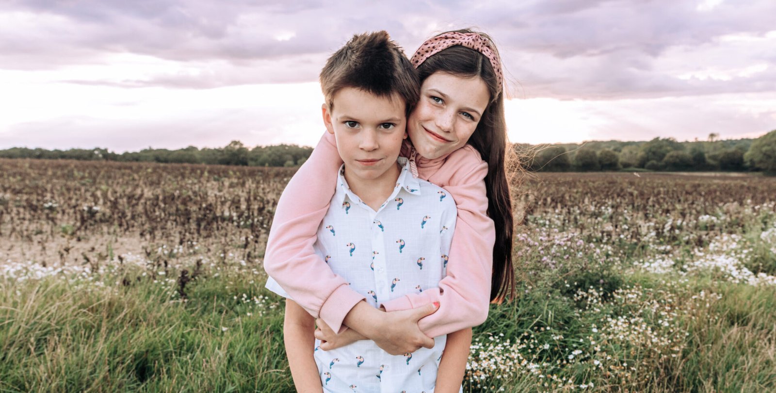 An older sister, hugging her brother from behind whilst they look into the camera, standing in a West Sussex field at sunset