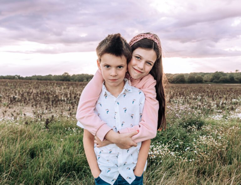 An older sister, hugging her brother from behind whilst they look into the camera, standing in a West Sussex field at sunset
