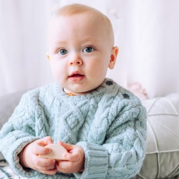 Sitting baby in the stuido, wearing a chunky mint-green jumper, looking into the camera with big blue eyes, with cushions behind him.